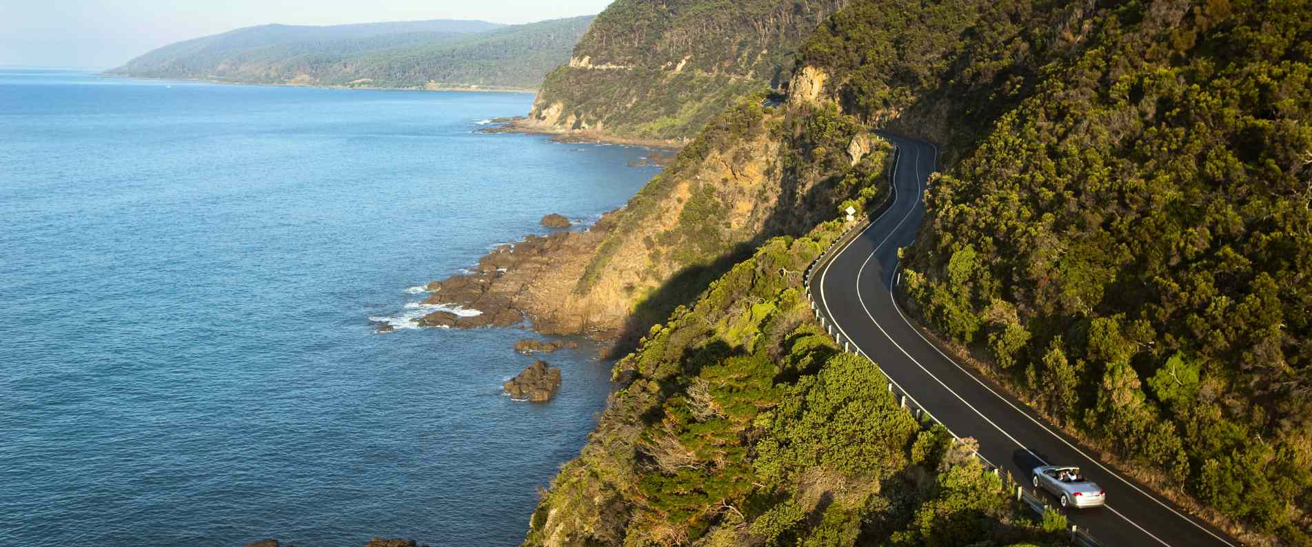 Aerial view of the winding Great Ocean Road curving along rugged cliffs with dramatic mountains meeting the ocean.
